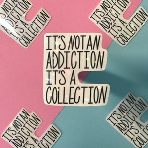 May include: White stickers with the text "IT'S NOT AN ADDICTION IT'S A COLLECTION" in black. The stickers have a holographic, glittery effect. The background is divided diagonally into pink and blue sections.