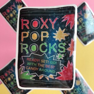 以下が含まれることがあります： 虹色の文字で「ROXY POP ROCKS」と書かれた黒と白のパッケージのステッカー。パッケージには赤、黄、緑の星形のデザインが施されています。「READY! SET! GO! WITH THE BEST CANDY AROUND」という文字が白い吹き出しの中に書かれています。
