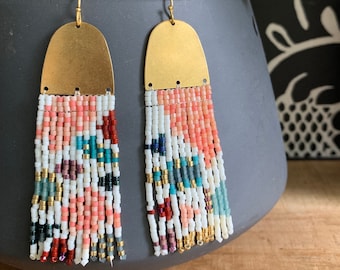 May include: A pair of handmade beaded earrings with a brass top and colorful fringe. The fringe features a pattern of white, coral, teal, and black beads. The earrings are displayed in front of a dark gray pot with a plant.
