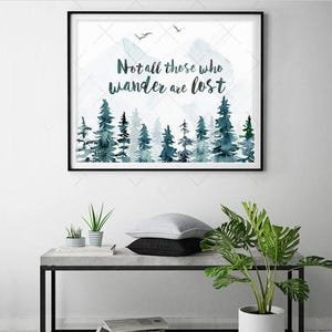 Può includere: Una stampa ad acquerello incorniciata con una citazione che recita "Not all those who wander are lost." La stampa presenta un'illustrazione ad acquerello blu e verde di una foresta con alberi.