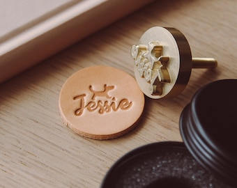 May include: A close-up of a leather stamp with the name "Jessie" and a small dog silhouette. The stamp is on a wooden surface next to a brass stamp with a design of a tree and the words "Just Breathe".
