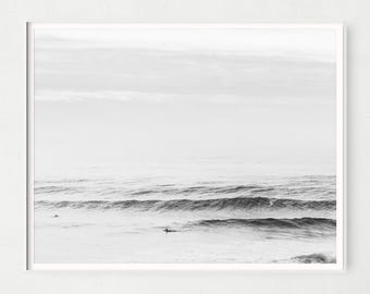 May include: Black and white framed print of ocean waves. The grayscale photograph captures the movement of the water with surfers in the distance. The sky is overcast, creating a soft, diffused light over the scene.