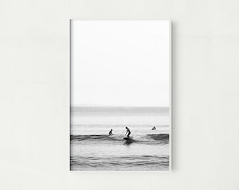 May include: Black and white framed print of surfers riding waves in the ocean. The image captures the silhouettes of surfers against the backdrop of the sea and sky, creating a minimalist aesthetic. The artwork is a perfect addition to any coastal-themed decor.