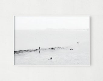May include: Black and white photograph of surfers in the ocean. The surfers are silhouetted against the water and the horizon. The photo is taken from a distance, so the surfers are small in the frame.