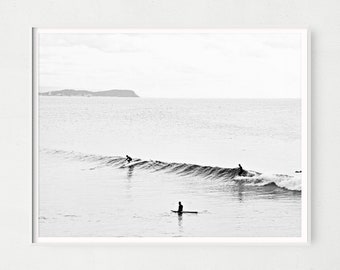 May include: Black and white photograph of three surfers riding waves in the ocean. The surfers are silhouetted against the water and sky. A distant island is visible in the background.