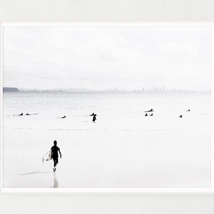 May include: A black and white photograph of a surfer walking on a beach with a surfboard under their arm. The ocean is in the background with a city skyline in the distance.