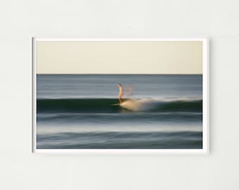May include: A blurry image of a person surfing on a wave in the ocean. The water is a light blue color and the sky is a light yellow color.