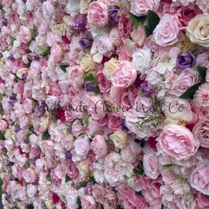 Puede incluir: Una pared cubierta de flores artificiales rosas, blancas y moradas. Las flores están dispuestas en una variedad de tamaños y formas, creando un aspecto exuberante y romántico.