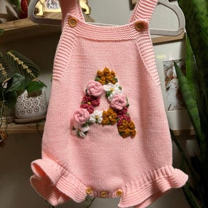 May include: Pink knitted baby romper with a floral "A" design. The romper features a ruffled hem, button closures, and shoulder straps with button accents. The floral design includes pink, white, and brown flowers.