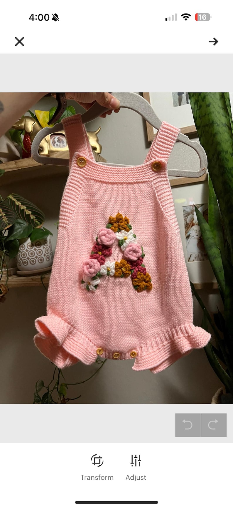 May include: Pink knitted baby romper with a floral "A" design. The romper features a ruffled hem, button closures, and shoulder straps with button accents. The floral design includes pink, white, and brown flowers.