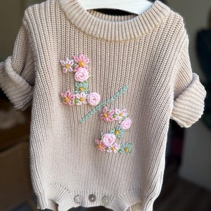 May include: A beige knit baby romper with short sleeves, featuring an embroidered floral design in pink, yellow, and green. The romper has a ribbed neckline and cuffs, and three buttons at the bottom. The garment is hanging on a white hanger.