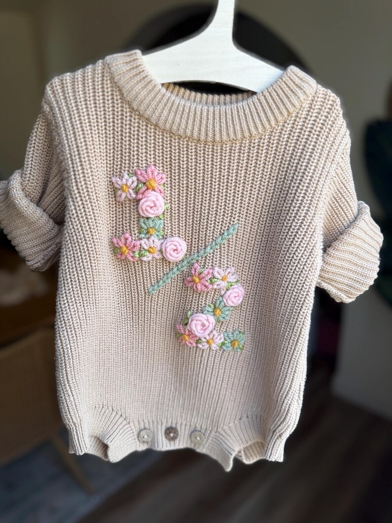 May include: A beige knit baby romper with short sleeves, featuring an embroidered floral design in pink, yellow, and green. The romper has a ribbed neckline and cuffs, and three buttons at the bottom. The garment is hanging on a white hanger.