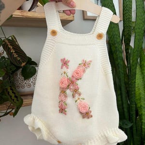 May include: Cream-colored knitted baby romper with a floral "K" design in pink and white. Features wooden buttons and a ruffled hem. The romper is displayed on a wooden hanger.