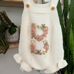 May include: Cream-colored knitted baby romper with a floral embroidered "B" on the front. The romper features wooden button closures and ruffled edges. A charming and personalized baby garment.