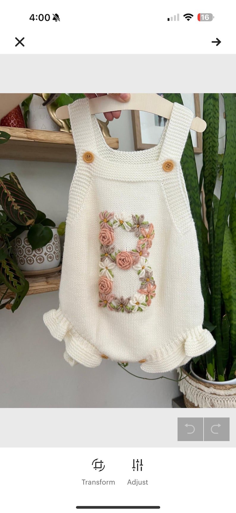 May include: Cream-colored knitted baby romper with a floral embroidered "B" on the front. The romper features wooden button closures and ruffled edges. A charming and personalized baby garment.