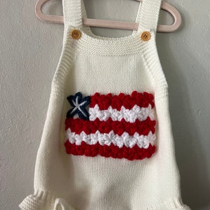 May include: White knitted baby romper with a crocheted American flag design on the front. The romper has ruffled edges and gold buttons.