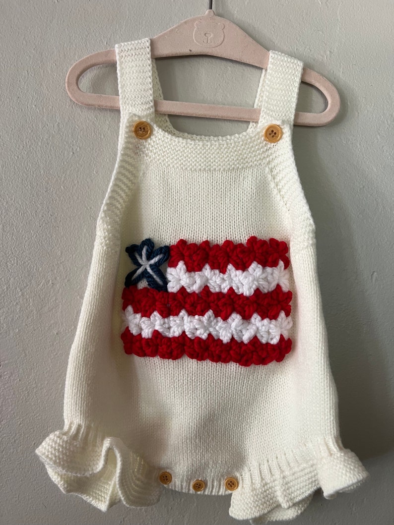 May include: White knitted baby romper with a crocheted American flag design on the front. The romper has ruffled edges and gold buttons.
