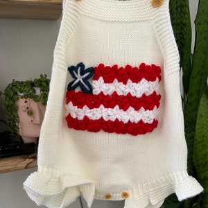 May include: White knitted baby romper with a red, white, and blue crocheted American flag design on the front. The romper has a ruffled hem and straps.