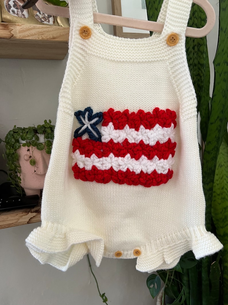May include: White knitted baby romper with a red, white, and blue crocheted American flag design on the front. The romper has a ruffled hem and straps.