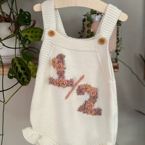 May include: Cream-colored knitted baby romper with floral embroidery spelling "1/2". The romper has wooden buttons on the straps and a ruffled hem. It is hanging on a wooden hanger.