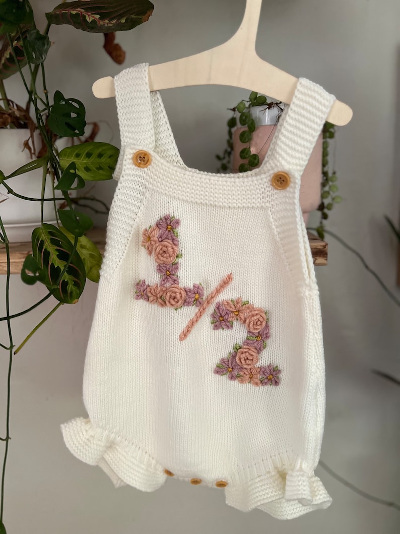May include: Cream-colored knitted baby romper with floral embroidery spelling "1/2". The romper has wooden buttons on the straps and a ruffled hem. It is hanging on a wooden hanger.