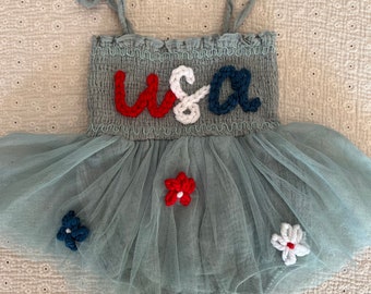 May include: A light blue tulle baby dress with a crocheted "USA" design on the bodice. The dress has red, white, and blue crocheted flowers on the skirt.