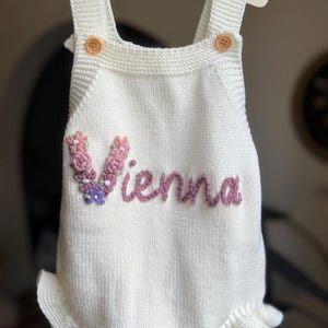 May include: White knitted baby romper with the name "Vienna" embroidered in pink. The romper features a floral embellishment and wooden buttons. The garment is displayed on a wooden hanger.