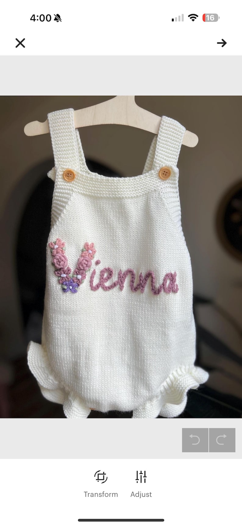 May include: White knitted baby romper with the name "Vienna" embroidered in pink. The romper features a floral embellishment and wooden buttons. The garment is displayed on a wooden hanger.