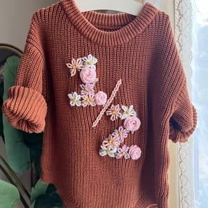 May include: A rust-colored knitted baby romper with short sleeves and wooden buttons. The romper features floral embroidery spelling out "1/2" in white, pink, and yellow. The garment is hanging on a white hanger.