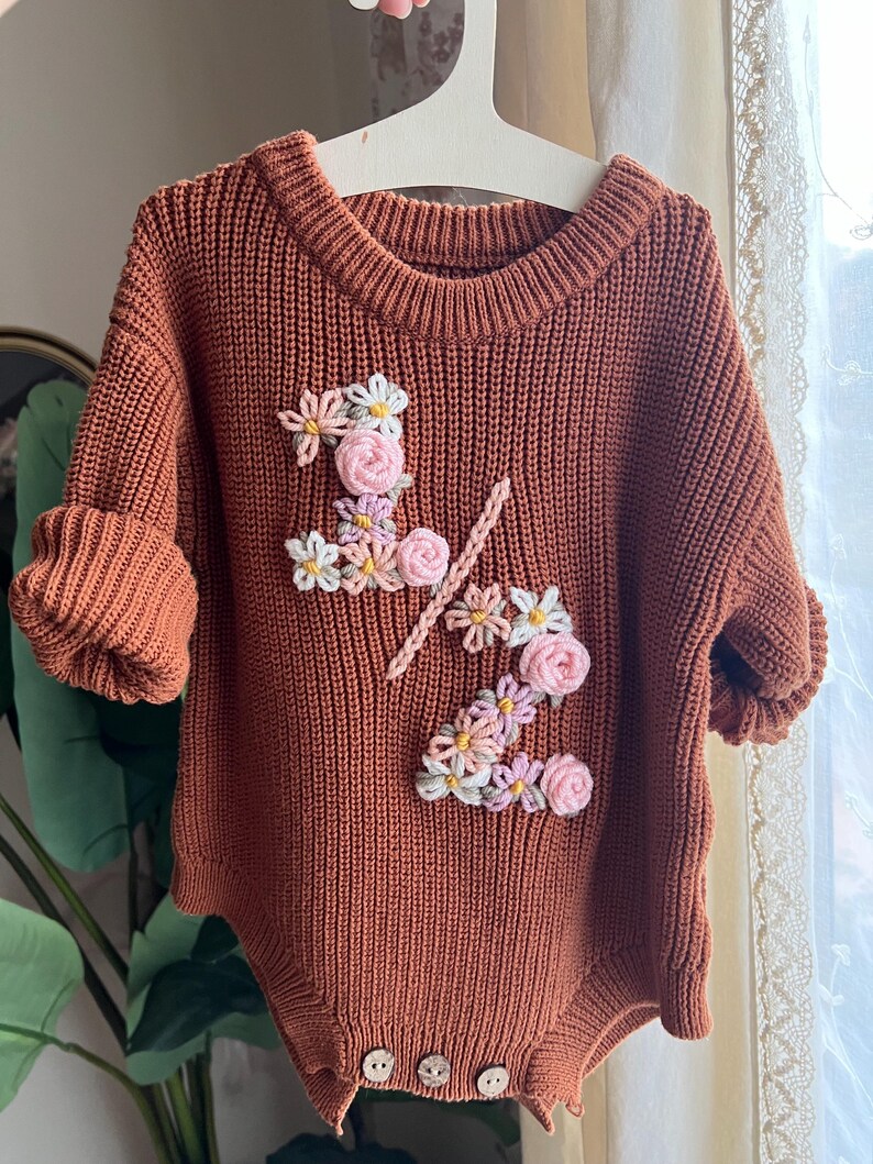 May include: A rust-colored knitted baby romper with short sleeves and wooden buttons. The romper features floral embroidery spelling out "1/2" in white, pink, and yellow. The garment is hanging on a white hanger.