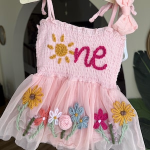 May include: A light pink sleeveless romper with a smocked bodice featuring an embroidered yellow sun and the word "one" in dark pink yarn. The skirt is made of layered tulle decorated with various colorful yarn-stitched flowers and stems.