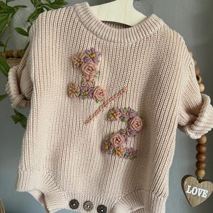 May include: A light pink knitted baby romper with the number "1 2" embroidered on the front with floral accents in pink, purple, and yellow. The romper has three brown buttons at the bottom and is hanging on a wooden hanger.