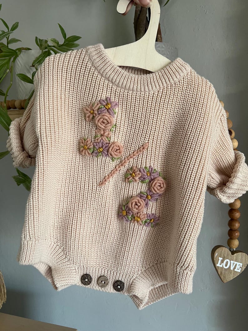 May include: A light pink knitted baby romper with the number "1 2" embroidered on the front with floral accents in pink, purple, and yellow. The romper has three brown buttons at the bottom and is hanging on a wooden hanger.