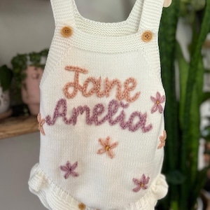 May include: White knitted baby romper with the name "Jane Amelia" embroidered in pink and purple thread. The romper features wooden buttons, floral accents, and a ruffled hem. The garment is displayed on a wooden hanger.