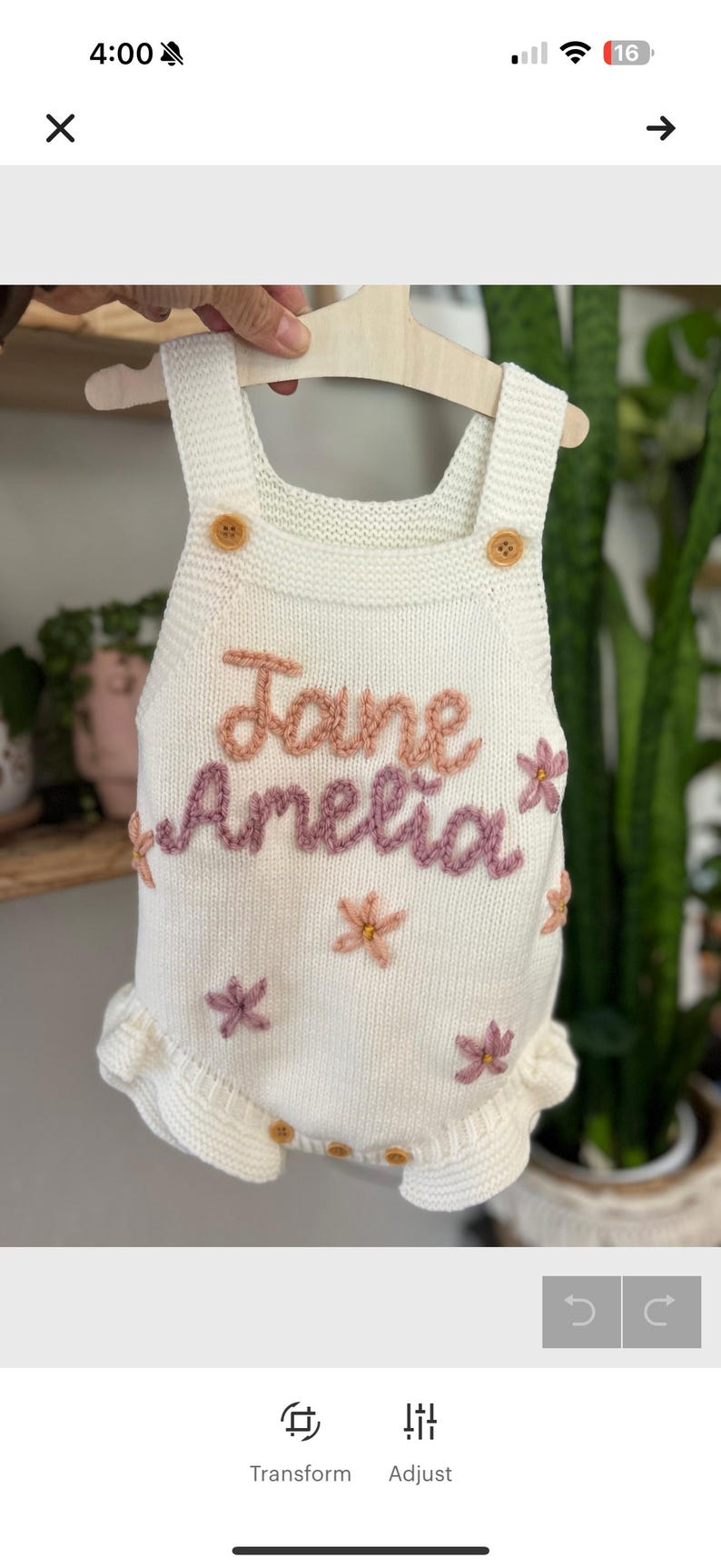 May include: White knitted baby romper with the name "Jane Amelia" embroidered in pink and purple thread. The romper features wooden buttons, floral accents, and a ruffled hem. The garment is displayed on a wooden hanger.
