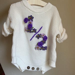 May include: A white knit romper with rolled sleeves, featuring the embroidered text "1/2" in purple and adorned with purple floral accents. The romper has three brown buttons at the bottom and is displayed on a wooden hanger.