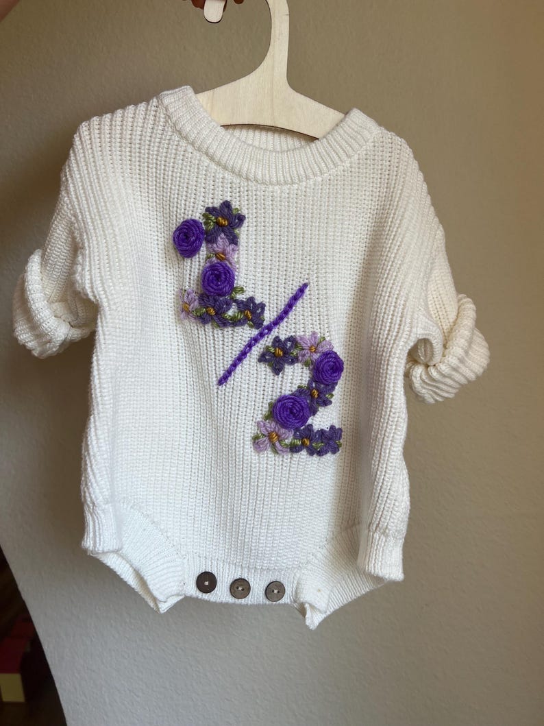 May include: A white knit romper with rolled sleeves, featuring the embroidered text "1/2" in purple and adorned with purple floral accents. The romper has three brown buttons at the bottom and is displayed on a wooden hanger.