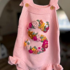 May include: A light pink knitted baby romper with a floral "S" design. The romper features button closures on the straps and a ruffled hem. The floral design includes pink, yellow, and orange flowers.