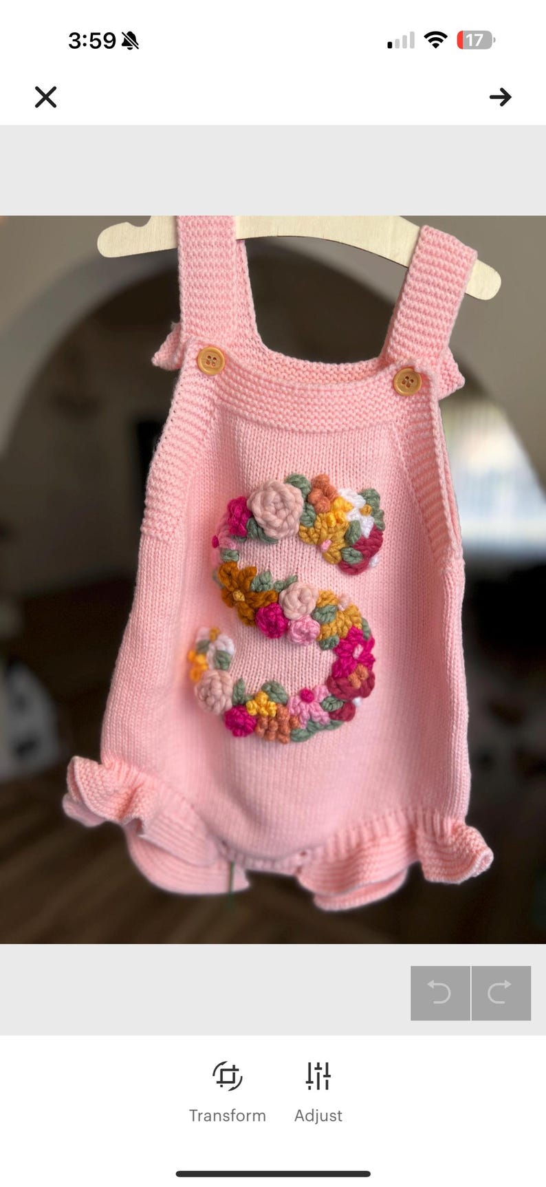 May include: A light pink knitted baby romper with a floral "S" design. The romper features button closures on the straps and a ruffled hem. The floral design includes pink, yellow, and orange flowers.