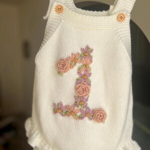 May include: Cream-colored knitted baby romper with a floral "1" design. The number is embroidered with pink and purple flowers and green leaves. Features include wooden buttons and a ruffled hem. The romper is displayed on a wooden hanger.