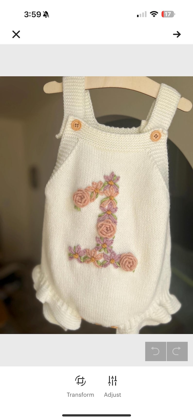 May include: Cream-colored knitted baby romper with a floral "1" design. The number is embroidered with pink and purple flowers and green leaves. Features include wooden buttons and a ruffled hem. The romper is displayed on a wooden hanger.