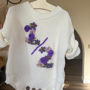 May include: White knit baby romper with purple floral embroidery forming the number "2" and a slash. The romper has short sleeves, wooden buttons, and is hanging on a wooden hanger. The design is likely for a baby's half-birthday.