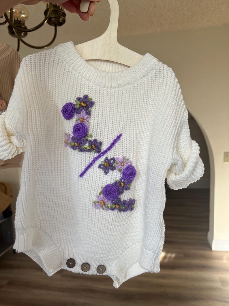 May include: White knit baby romper with purple floral embroidery forming the number "2" and a slash. The romper has short sleeves, wooden buttons, and is hanging on a wooden hanger. The design is likely for a baby's half-birthday.