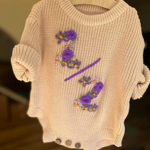 May include: A light beige knitted baby romper with short sleeves, featuring embroidered purple floral details forming the number "2/2". The romper has three wooden buttons at the bottom and is displayed on a wooden hanger.