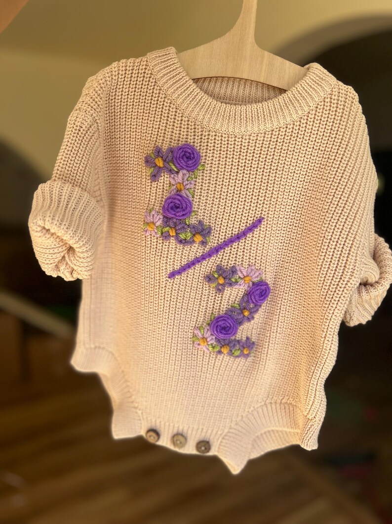 May include: A light beige knitted baby romper with short sleeves, featuring embroidered purple floral details forming the number "2/2". The romper has three wooden buttons at the bottom and is displayed on a wooden hanger.