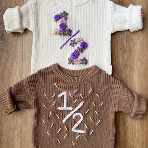 May include: Two baby outfits: a cream-colored sweater with purple floral embroidery forming "1/2", and a brown romper with white embroidered "1/2". Both are knit, with long sleeves, and are displayed on a wooden surface.