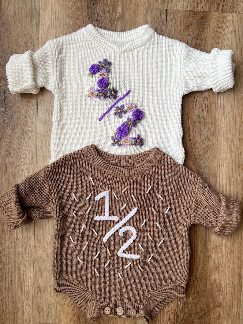 May include: Two baby outfits: a cream-colored sweater with purple floral embroidery forming "1/2", and a brown romper with white embroidered "1/2". Both are knit, with long sleeves, and are displayed on a wooden surface.