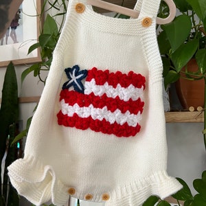 May include: White knitted baby romper with a red, white, and blue crocheted American flag design.
