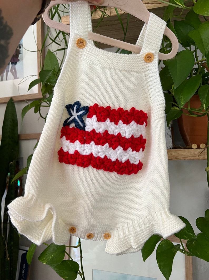 May include: White knitted baby romper with a red, white, and blue crocheted American flag design.