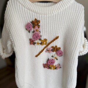 May include: A white knit baby bodysuit with short sleeves, featuring embroidered floral accents in pink, yellow, and brown. The bodysuit has a round neckline and three wooden buttons at the bottom. The garment is hanging on a wooden hanger.